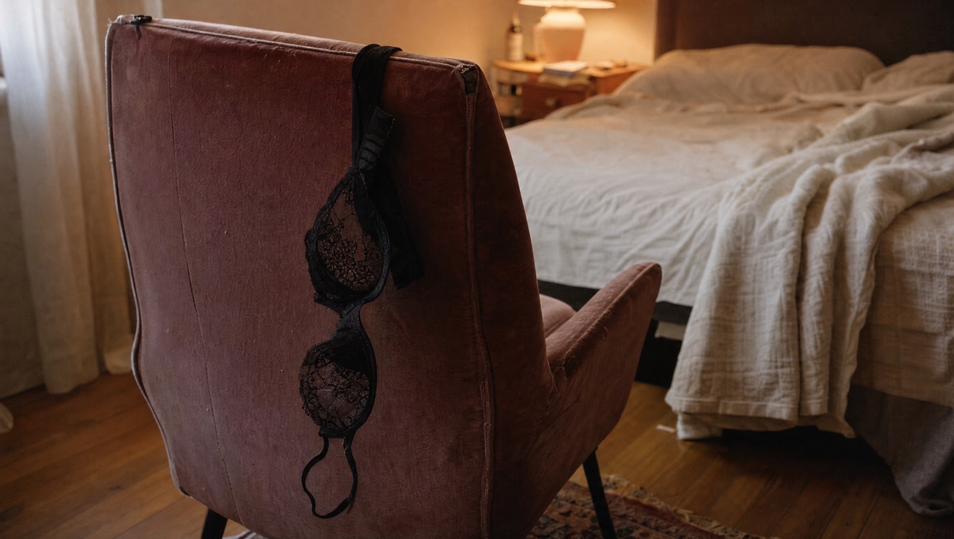 Black lace bra hanging over the back of a rose velvet chair in a dim bedroom