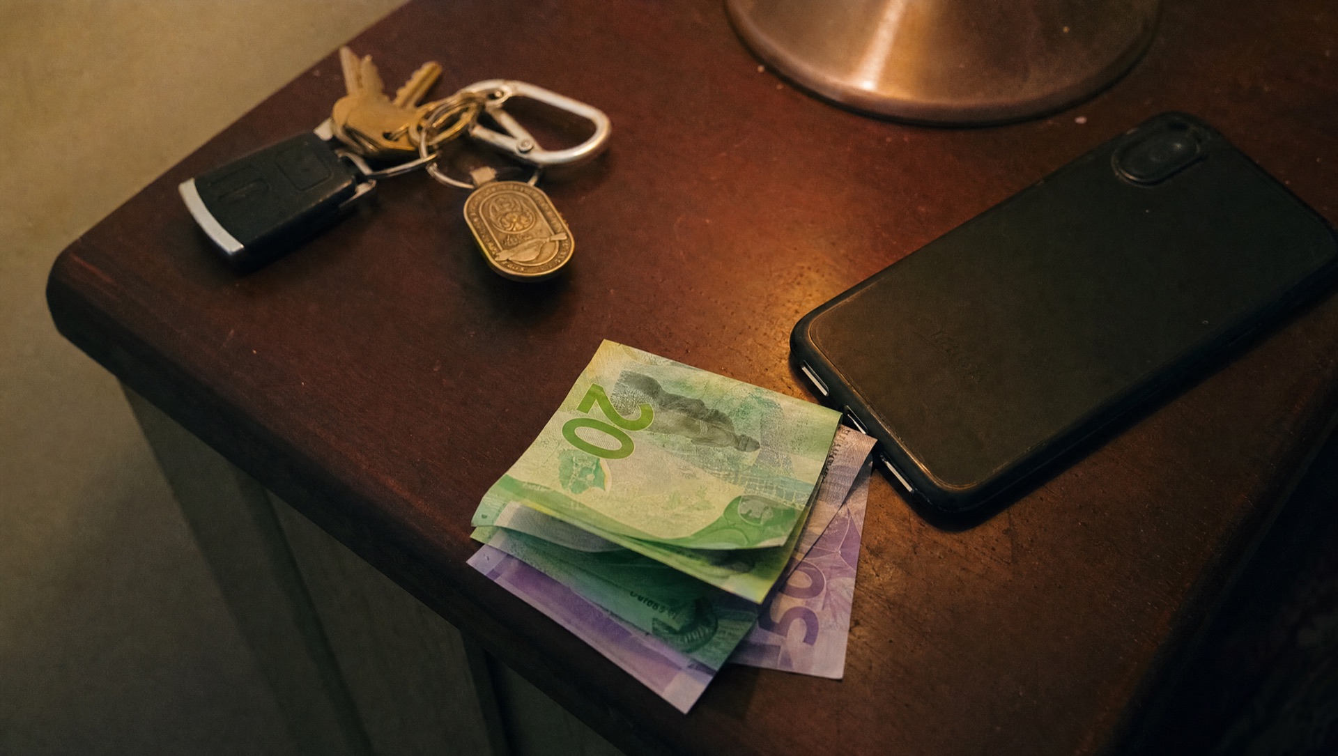 Folded New Zealand banknotes on a bedside table next to keys and a phone under lamp light