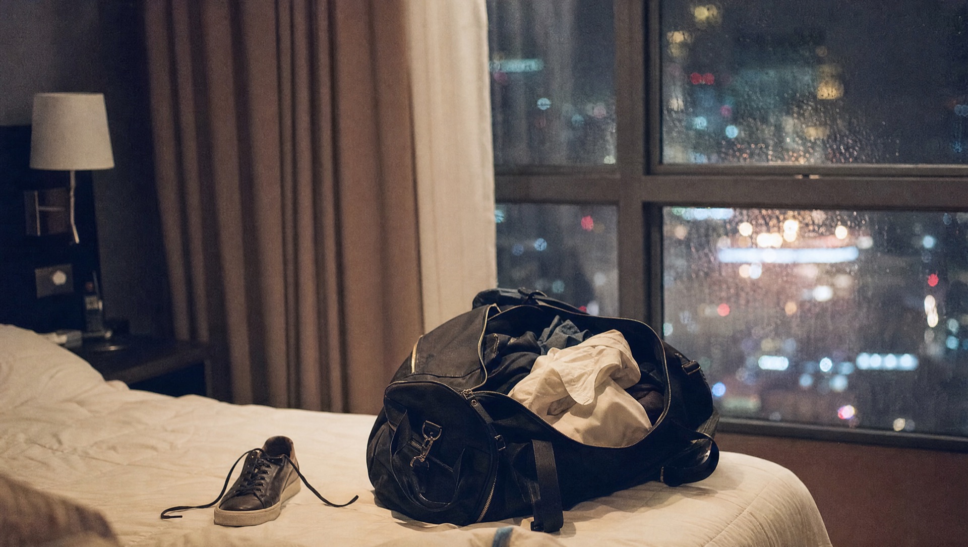 Half-packed overnight bag on a hotel bed with city lights through a rainy window at night